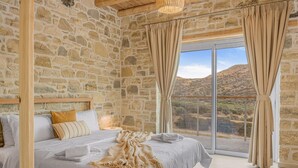 A bedroom with a stone wall, a large bed, beige curtains, and a view of a mountainous landscape. - Villa Nala With Pool by Matala (Faistos)