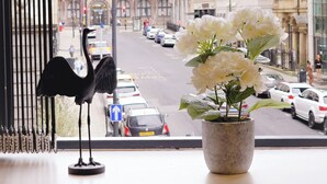 Une fenêtre donnant sur une rue, un pot de fleurs avec des fleurs blanches et une statuette d’oiseau noir.