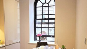 Un petit coin repas avec une table ronde dressée pour deux personnes, une fenêtre donnant sur des immeubles et un meuble placé sur le côté gauche.