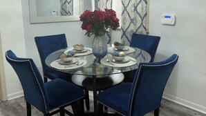 Een eetgedeelte met een ronde glazen tafel, blauwe fauteuils, een vaas met rode bloemen en een hanglamp.