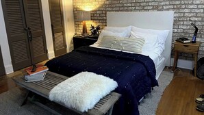 Ein Schlafzimmer mit einer Ziegelwand, einem Bett mit weißer und dunkelblauer Bettwäsche, einem Nachttisch mit Lampe und einer hölzernen Bank mit weißem Kissen.