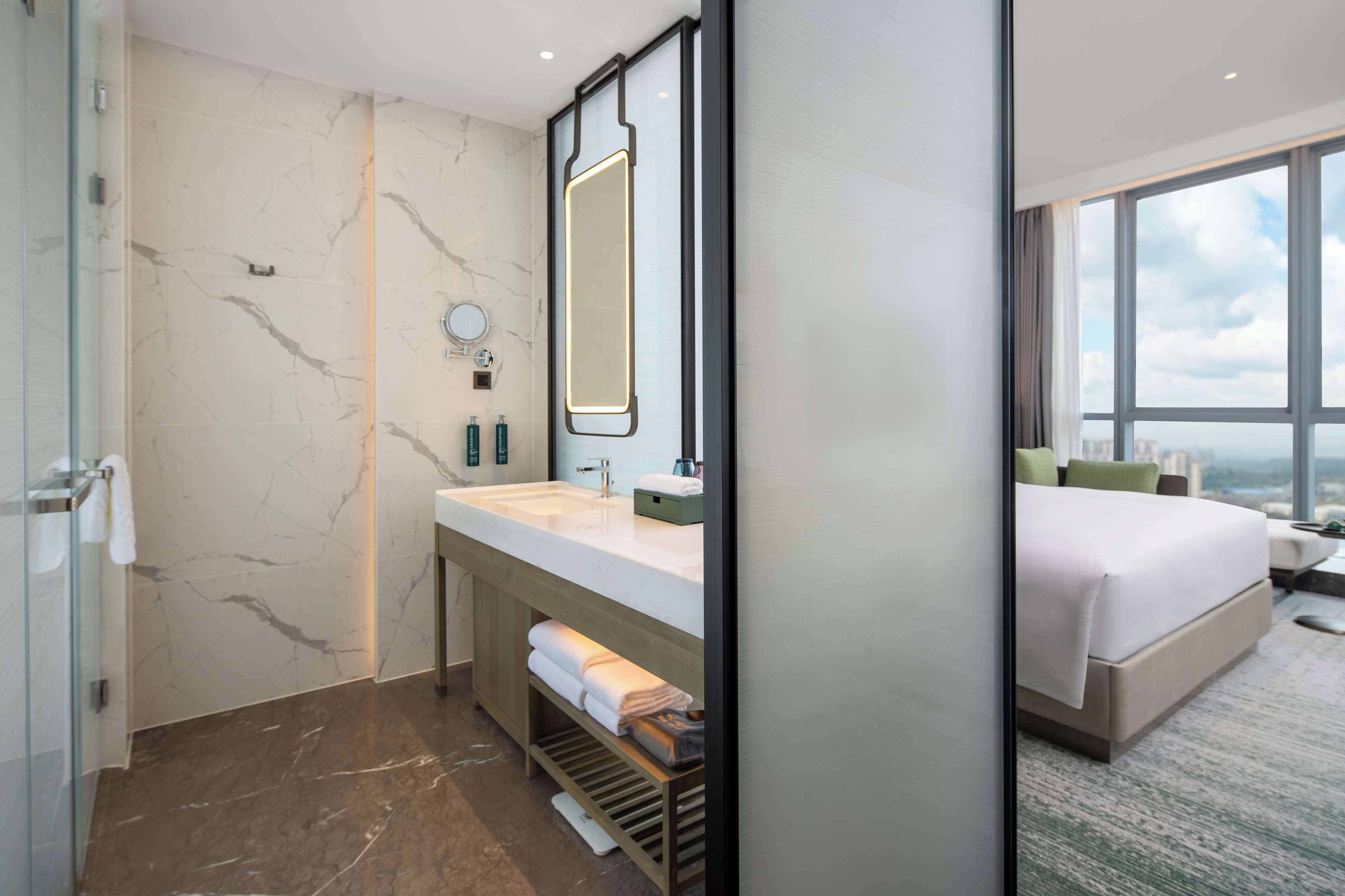 A modern hotel bathroom with a large mirror, marble walls, a sink, and a shower.