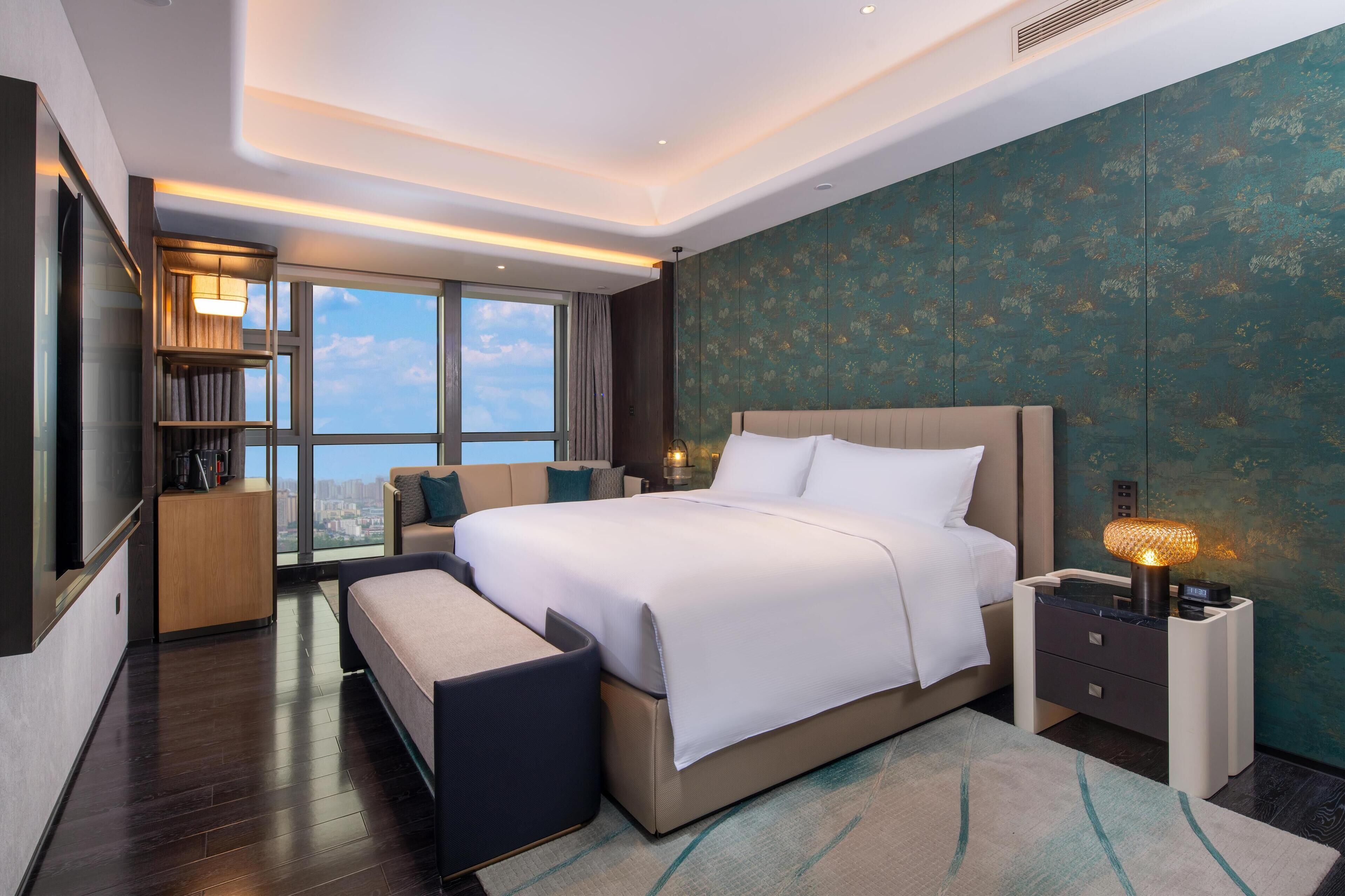 Una camera d'hotel moderna con un letto grande, un comodino con una lampada, una panca, una TV e vista sulla città attraverso ampie finestre.