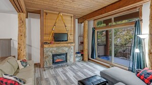 A cozy living room with a stone fireplace, wooden beams, a television mounted above it, and a large sliding glass door to the outdoors. - Champagne Powder Chalet! Best Ski In/Out at Big White. Sleeps up to 7ppl. (Beaverdell)