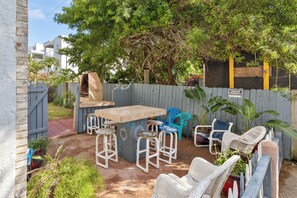 Ein Garten mit einem Holztisch und Barhocker, umgeben von Korbstühlen und einem blauen Plastikstuhl.