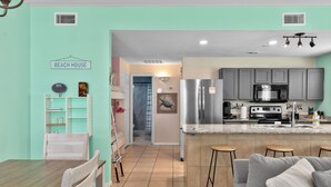 Una cocina moderna con una isla central, armarios grises y electrodomésticos de acero inoxidable. Cuenta con una zona de comedor con una mesa y sillas de madera, y un salón con un sofá gris.