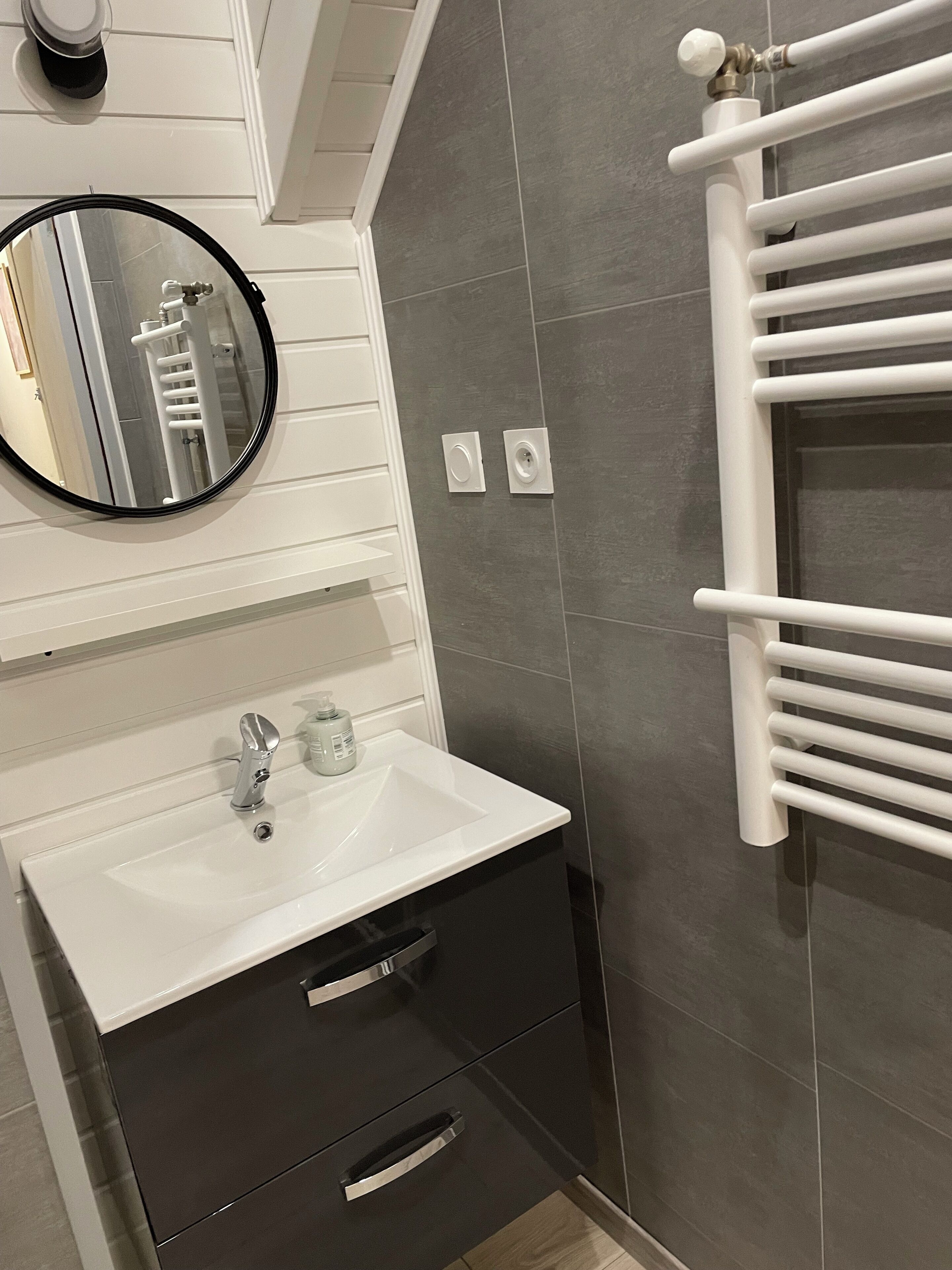Une salle de bain avec un lavabo blanc, des meubles de rangement aux couleurs sombres, un miroir rond et un porte-serviettes blanc.