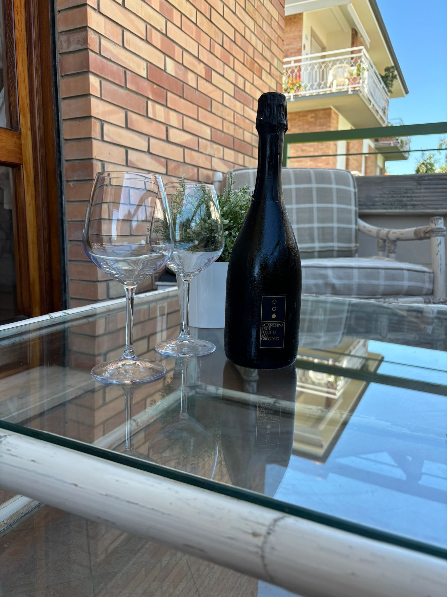A glass table with two wine glasses and a bottle of Falanghina Brun Brut di Falò di Gregorio wine.