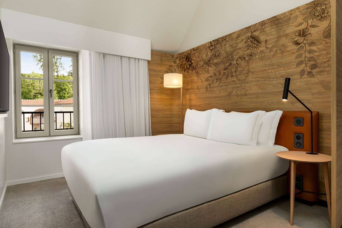 A hotel room with a large bed, a bedside table with a lamp, a window with curtains, and a wooden wall with floral patterns.