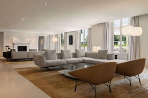 A modern living room with a sectional sofa, a coffee table, and two armchairs. There is a fireplace, a television, and large windows with curtains.