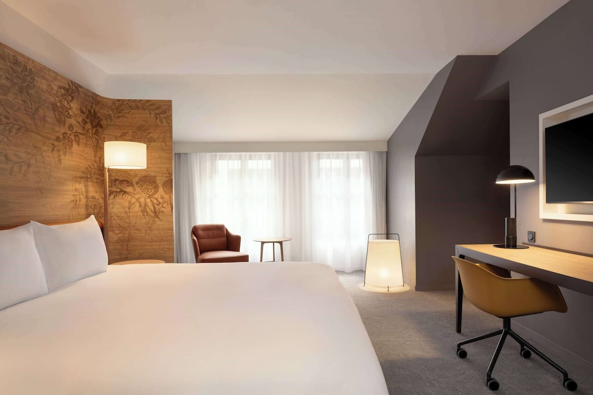 A hotel room with a large bed, a desk with a chair, a small table, and a lamp.