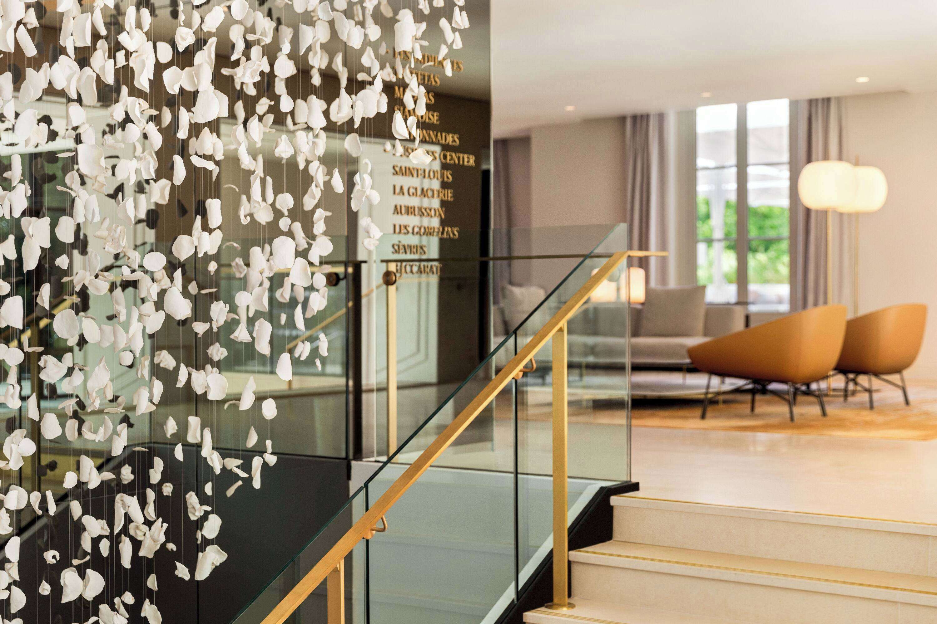 Un hall d’hôtel moderne doté d’un escalier en verre, d’un espace salon avec des fauteuils contemporains et d’un mur décoratif agrémenté d’éléments suspendus.