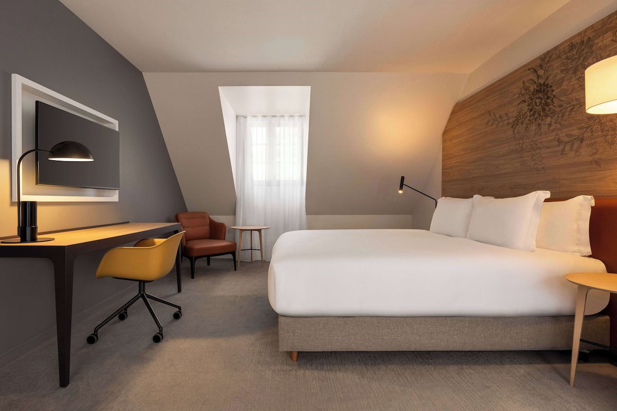 A modern hotel room with a large bed, a desk with a chair, a small table, and a decorative headboard.