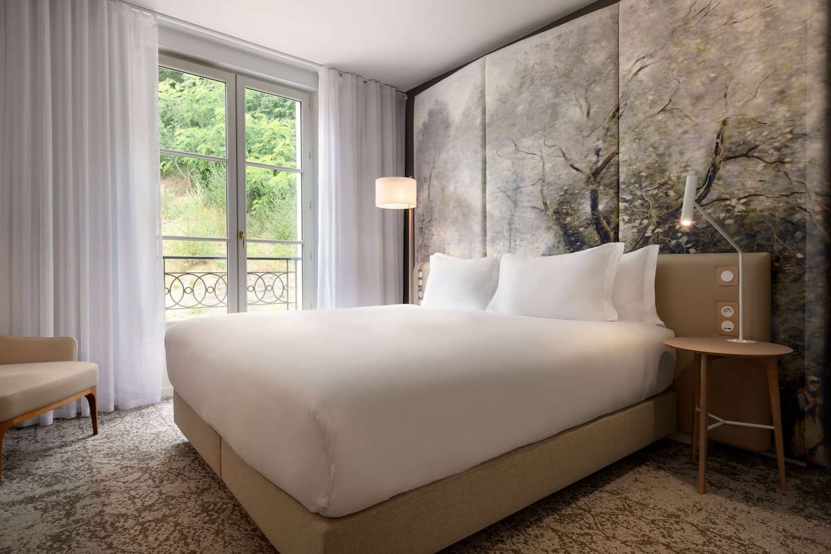 A hotel room with a large bed, a bedside table, a lamp, and a view of greenery through the window.