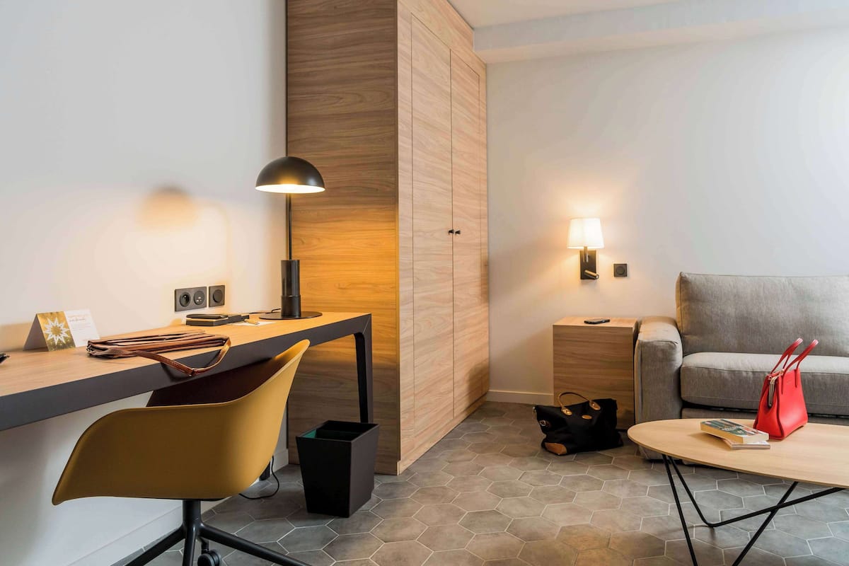 A modern hotel room with a wooden accent wall, a desk with a lamp, a sofa, and a coffee table.