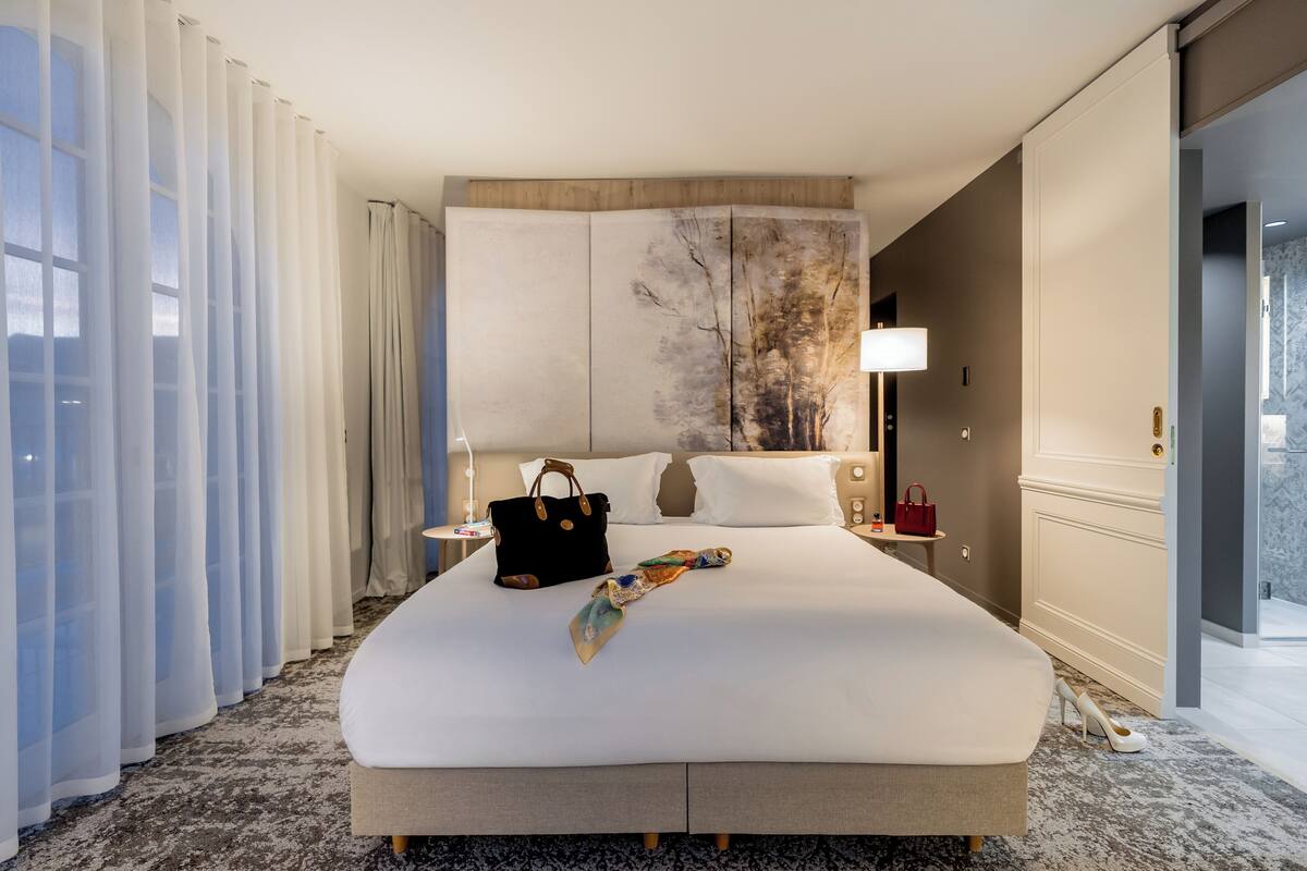 A hotel room with a large bed, a headboard with a painting, a nightstand with a lamp, and a view of the outside through the window.