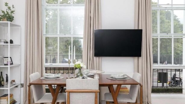 Een eetgedeelte met een houten tafel en stoelen, een boekenkast en een groot raam met gordijnen.