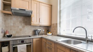 A modern kitchen with wooden cabinets, a stainless steel oven, a built-in range hood, a granite countertop, a sink, and a window with blinds. - Charming London City Apartment (London)