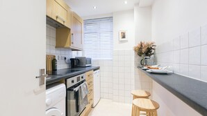 Une cuisine compacte équipée d’un lave-linge, d’un four à micro-ondes et d’une machine à café. On y trouve deux tabourets en bois et un vase avec des fleurs posé sur le comptoir.