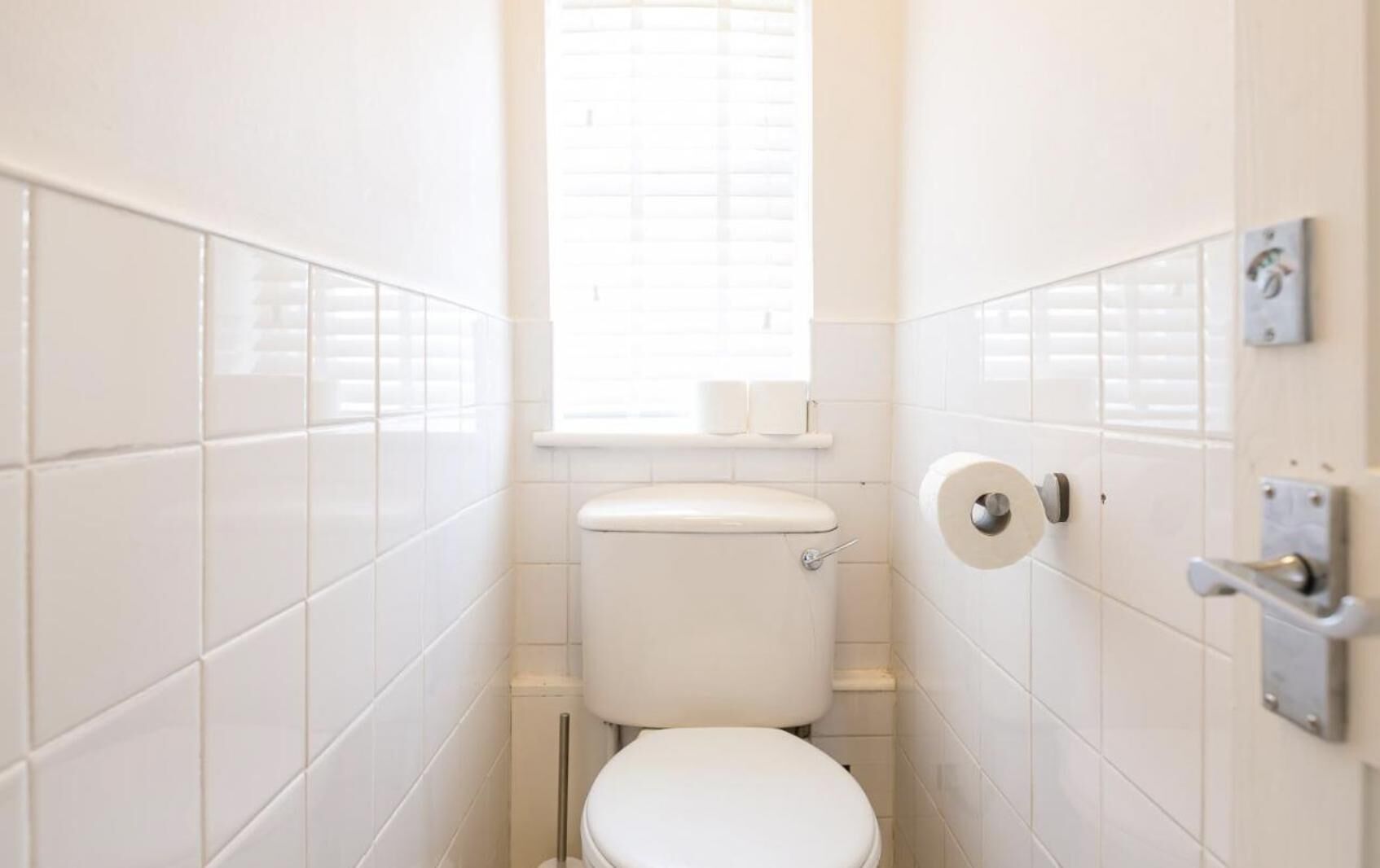 Une petite salle de bain aux murs recouverts de carreaux blancs, équipée de toilettes, d’une fenêtre et d’un porte-rouleau de papier toilette.