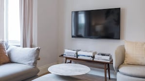 Ein Wohnzimmer mit Flachbildfernseher, Couchtisch und einem Stapel Bücher auf einem Holznachttisch.