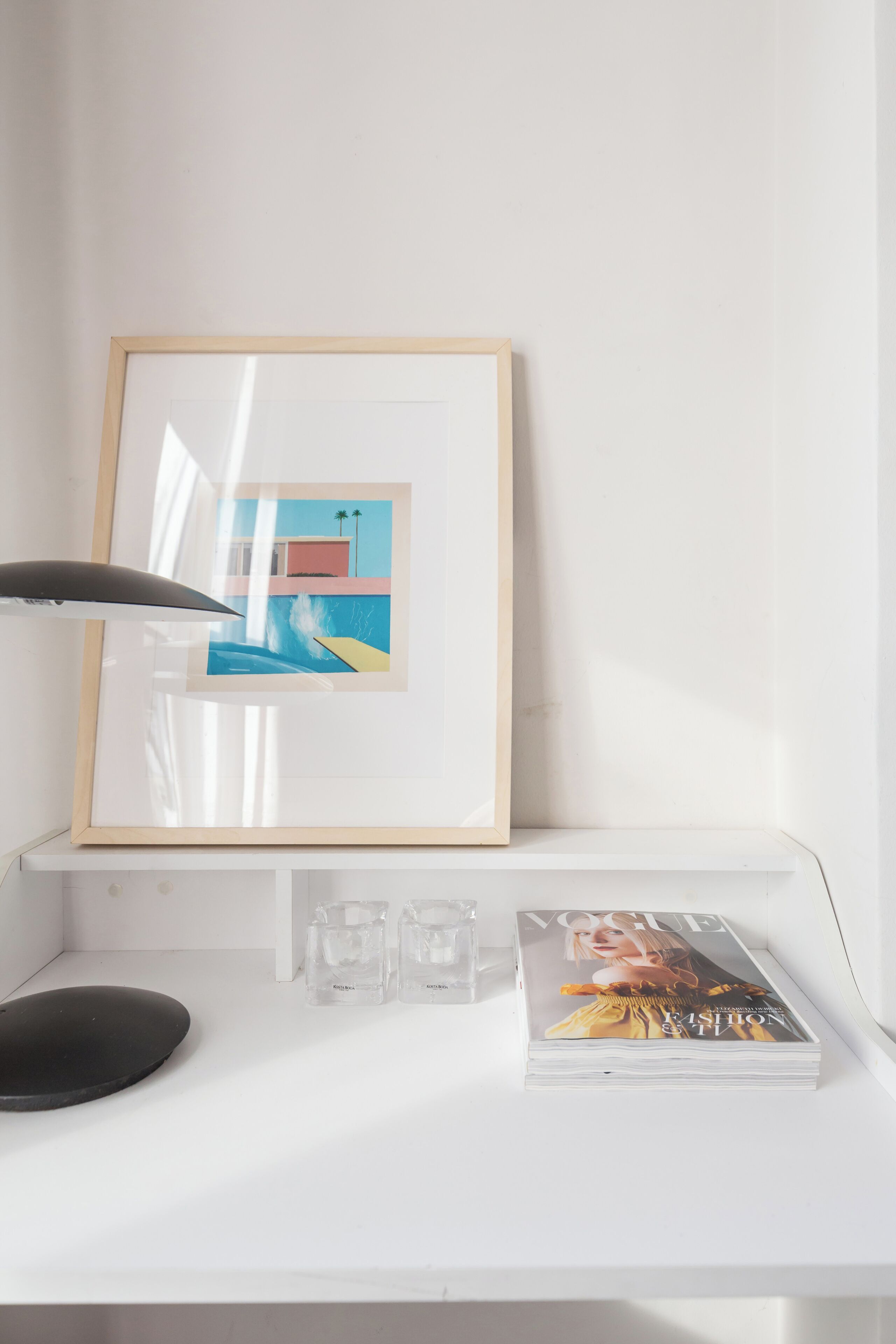 Een wit bureau met een lijst, een stapel tijdschriften en een zwarte lamp.