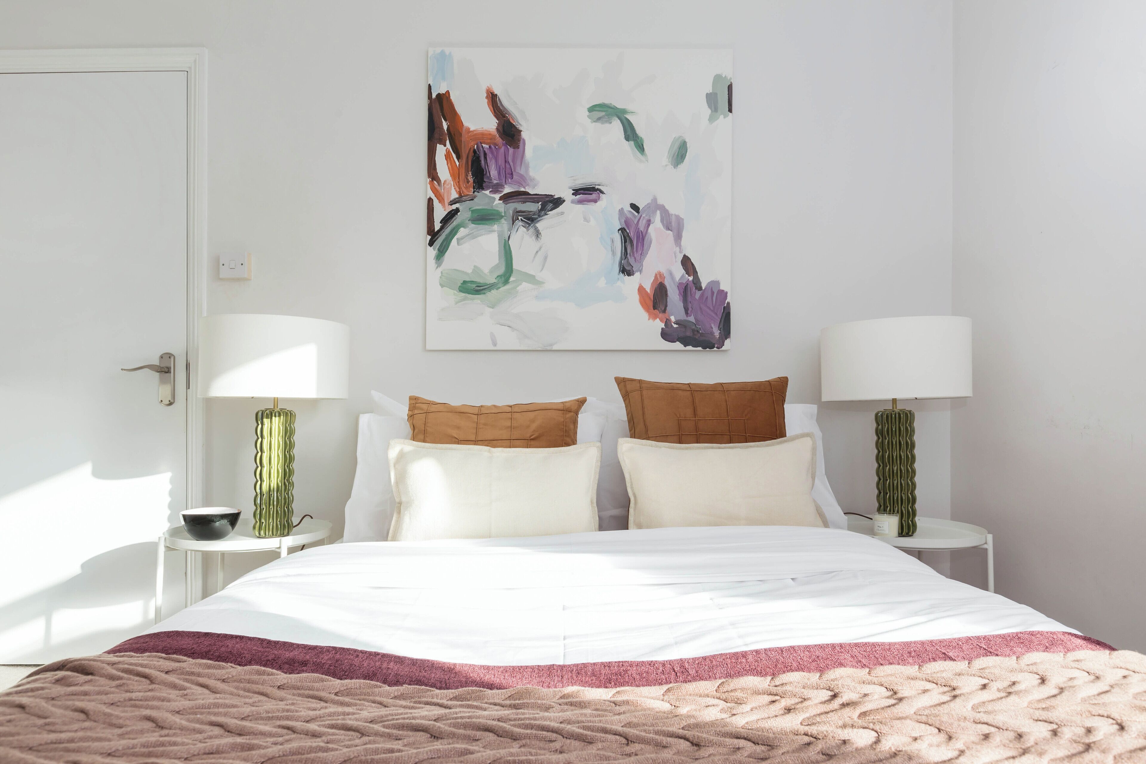 Une chambre à coucher comprenant un lit, deux tables de chevet, deux lampes, une œuvre d’art murale et une porte.