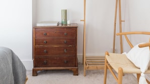 Ein Zimmer mit einem hölzernen Schminktisch, einem Holzstuhl mit gewebtem Sitz und einem Wäscheständer aus Holz vor einer weißen Wand.
