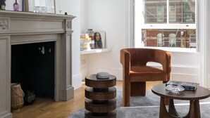 A modern living room with a fireplace, a large window, a brown armchair, and a round coffee table. - Generously Large 1 Bedroom Apartment in Farringdon (London)