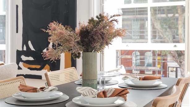 A dining area with a dark wooden table set for four, surrounded by woven chairs. A tall vase with dried flowers is placed on the table. - Generously Large 1 Bedroom Apartment in Farringdon (London)