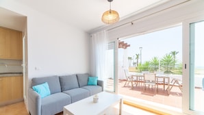 Un salon moderne avec un canapé gris, une table basse blanche et une grande porte-fenêtre coulissante donnant sur une terrasse extérieure équipée d’une table et de chaises.