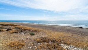 Une plage avec de l’herbe sèche et des galets, qui descend vers l’océan.