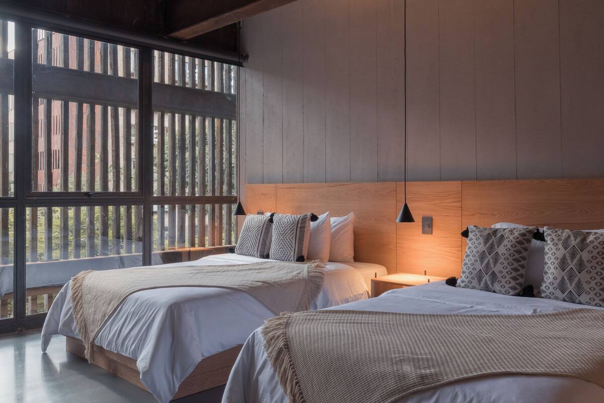 A bedroom with two beds, a wooden headboard, a hanging pendant light, and a large window with vertical blinds.