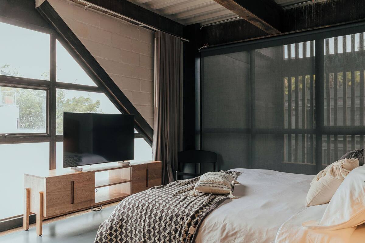 A bedroom with a large bed, a TV on a wooden cabinet, and a window with curtains.