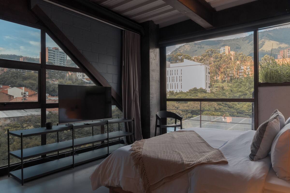 A bedroom with a large window, a bed, a TV on a stand, and a chair.