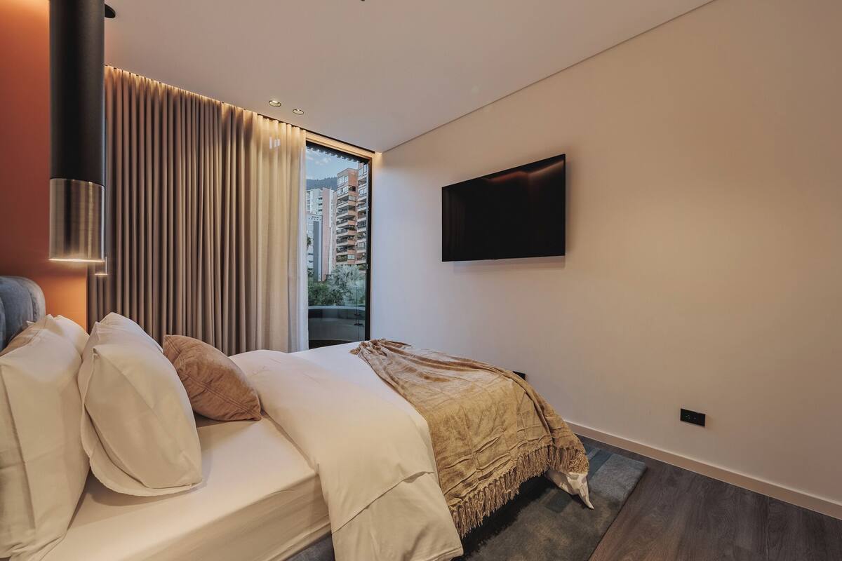 A modern hotel room with a bed, a large window with curtains, a flat-screen TV mounted on the wall, and a small rug on the floor.