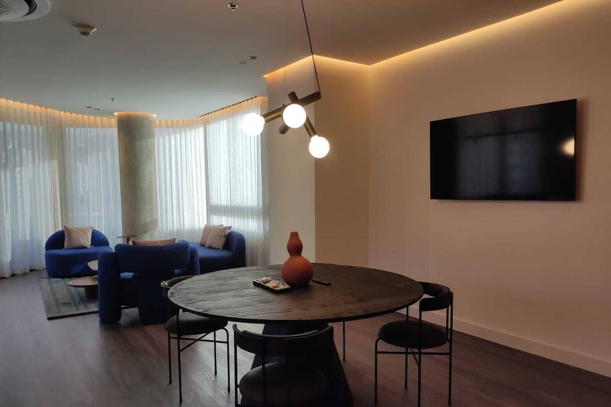 A modern living room with a round wooden table, blue sofas, and a large flat-screen TV mounted on the wall.