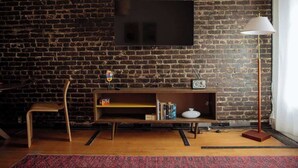 Een woonkamer met een bakstenen muur, een flatscreen televisie boven een houten dressoir, een houten stoel en een vloerlamp.
