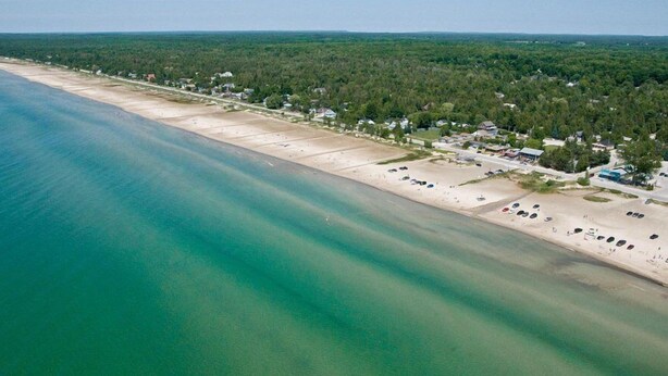 An aerial view of a coastal area with a sandy beach, a row of houses, and a dense forest in the background. - The Sands (Sauble Beach)