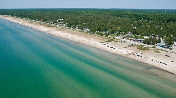 An aerial view of a coastal area with a sandy beach, a row of houses, and a dense forest in the background. - The Sands (Sauble Beach)