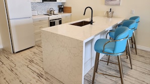 A modern kitchen with white cabinetry, a marble countertop island, a sink, and blue bar stools. - The Sands (Sauble Beach)