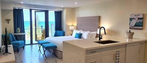 A hotel room with a bed, a small seating area, a kitchen island, and a view of the ocean.