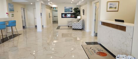 A hotel lobby with a reception desk, a seating area with patterned sofas, a bar with blue chairs, and a marble floor.
