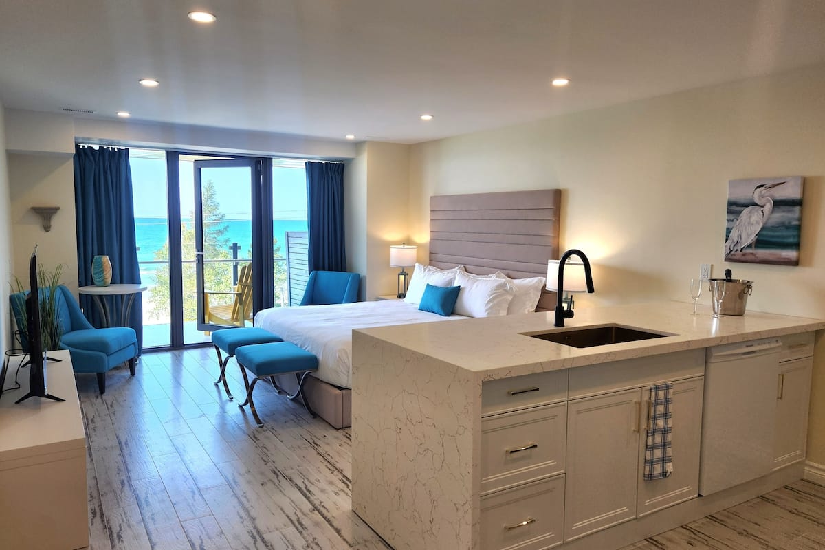 A modern hotel room with a large bed, a sitting area with a view, a kitchen with a sink, and a balcony with a view of the sea.