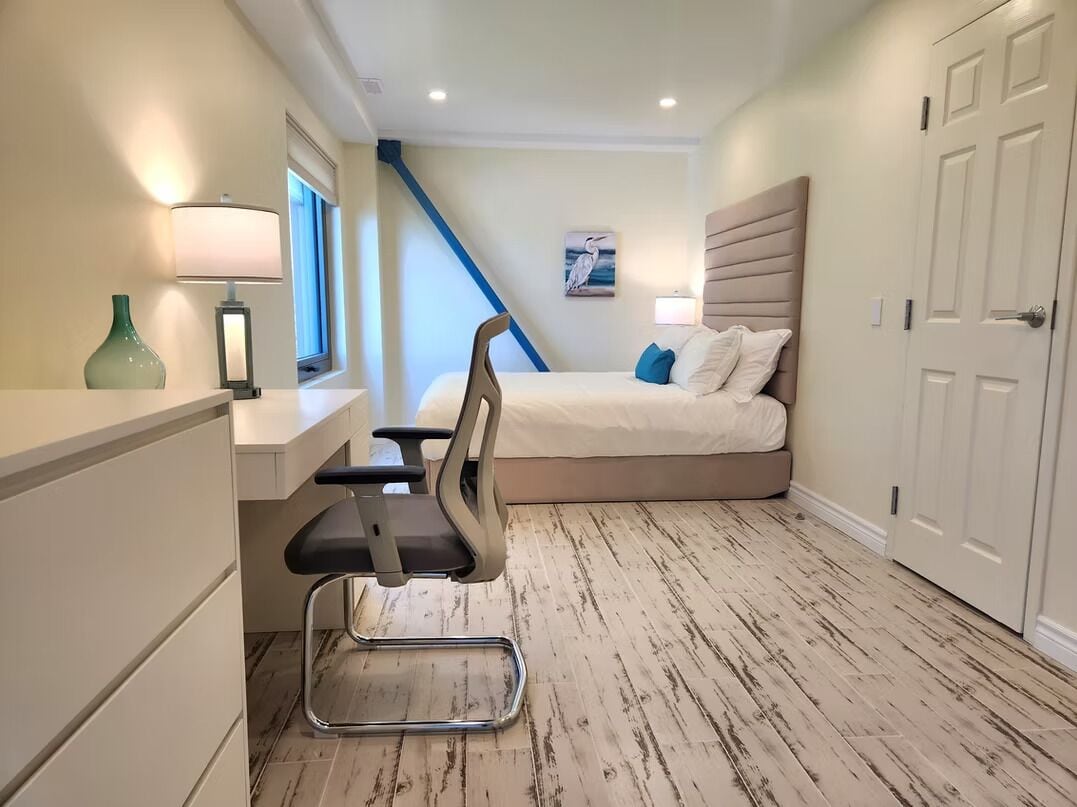Un dormitorio con cama, mesita de noche, lámpara, silla y vista al ventanal.