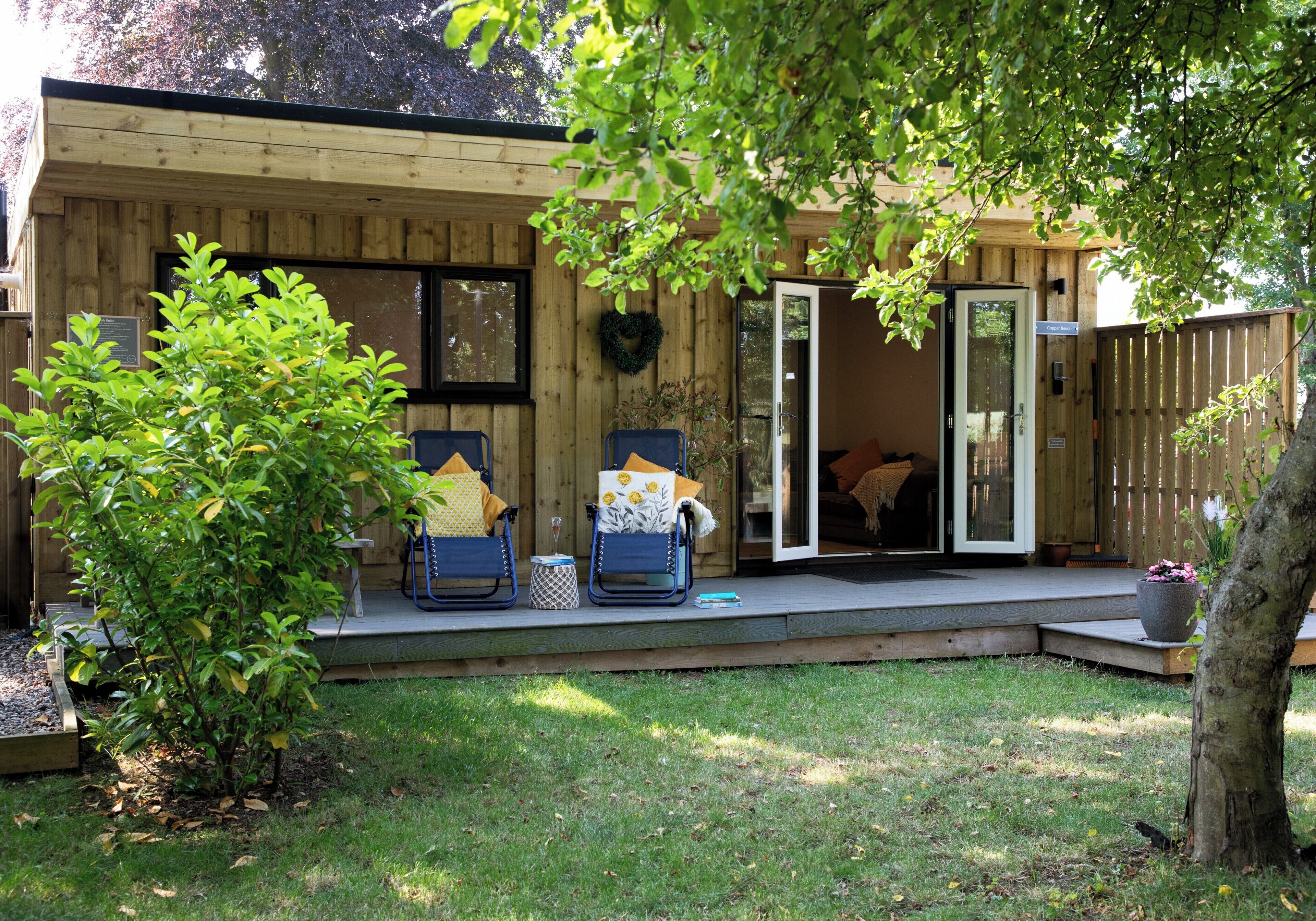 A wooden cabin with a covered patio, two lounge chairs, a small table, and a potted plant.