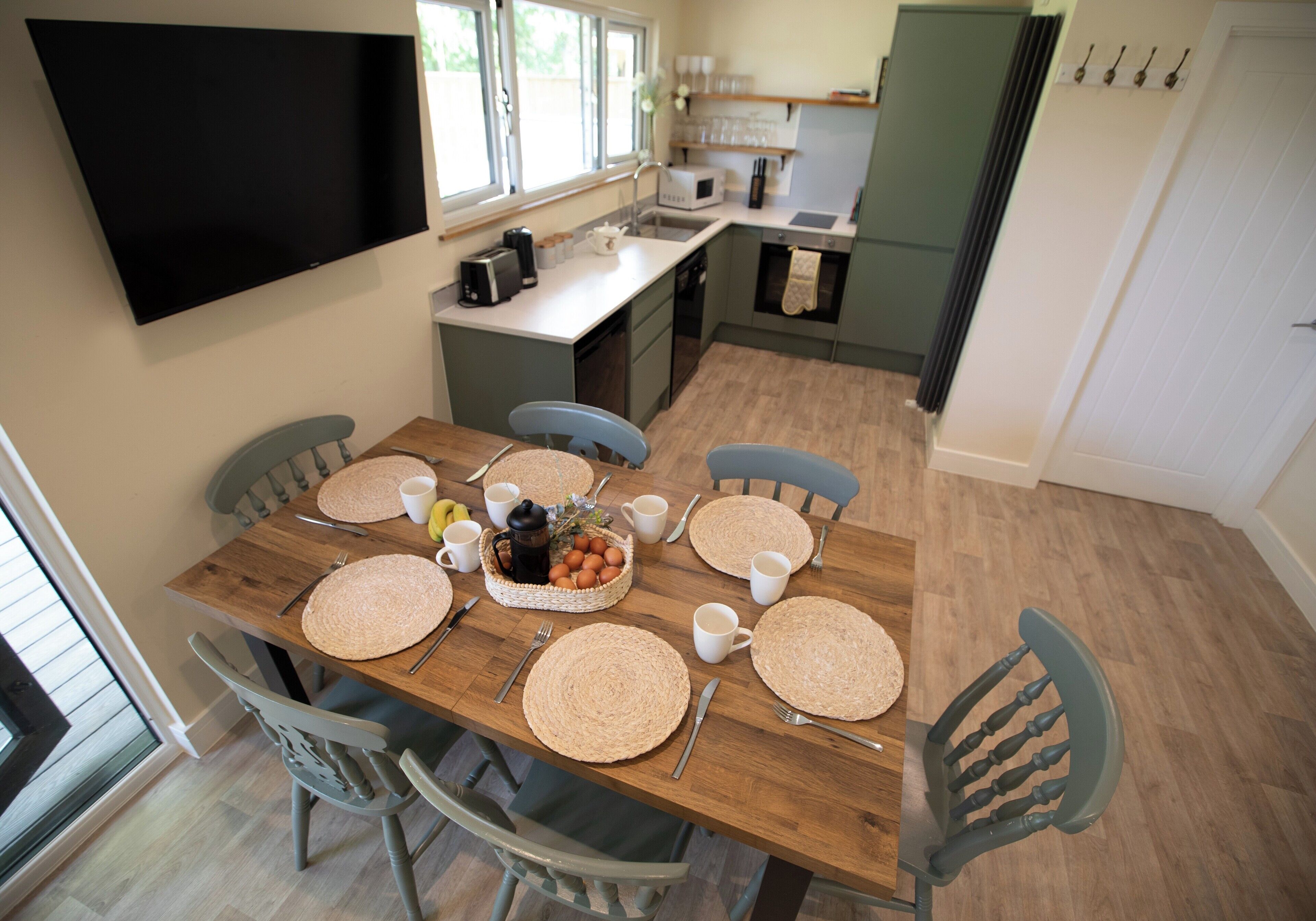A dining area with a wooden table set for six, surrounded by six chairs. A flat-screen TV is mounted on the wall above the table.