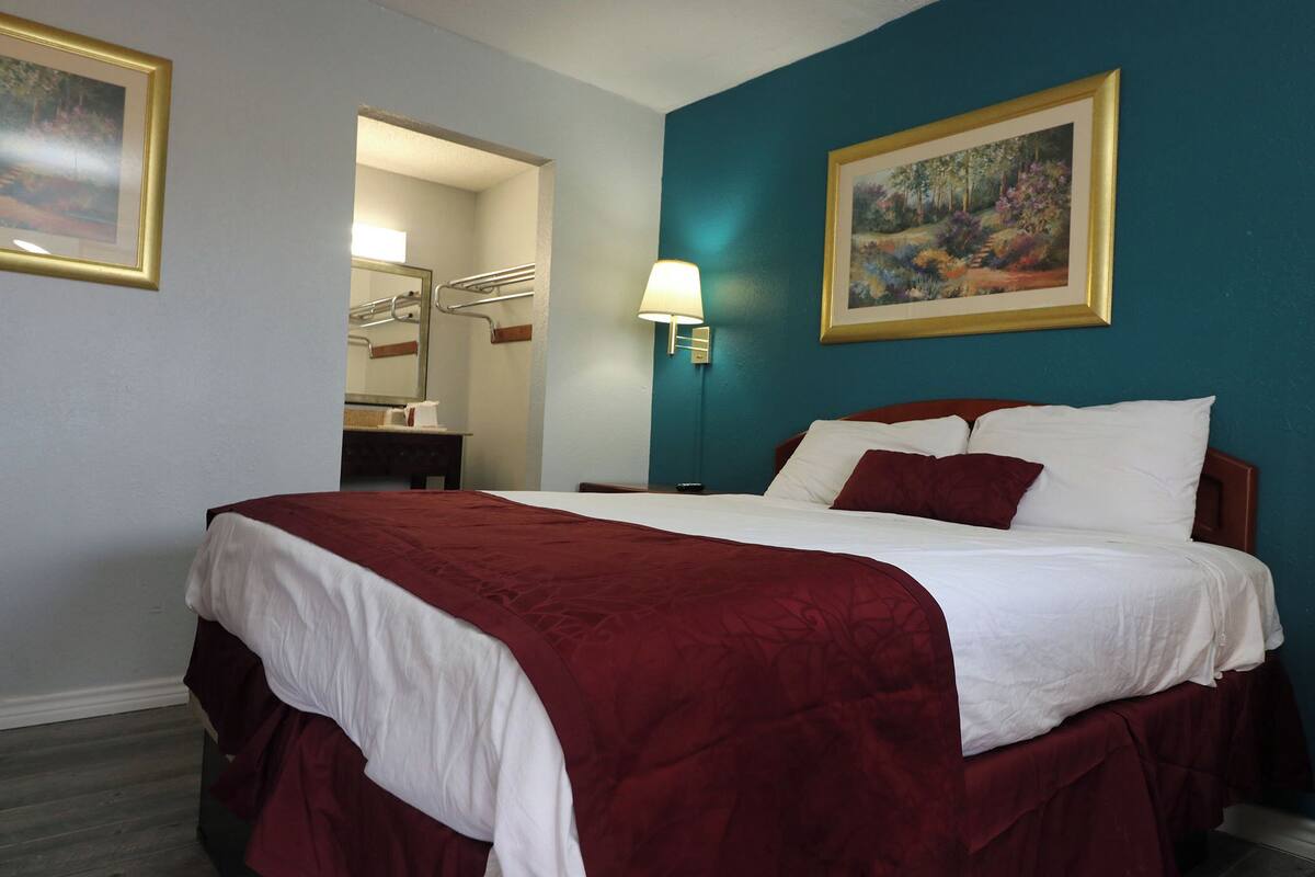 A hotel room with a large bed, a nightstand, a lamp, a painting on the wall, and a bathroom visible through an open door.