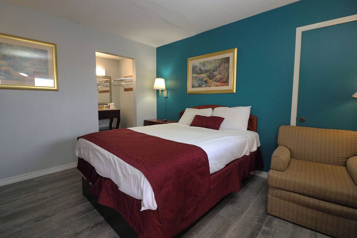 A hotel room with a large bed, a chair, a desk, and a painting on the wall.