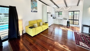A spacious living area with a yellow sofa, wooden flooring, a ceiling fan, and a kitchen with white cabinets. - Garden Retreat with Pool - fully detached Studio (Freshwater)